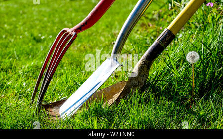 Eine rote Gartenarbeit Gartenarbeit Gabel einen Spaten und eine Gartenbau Schaufel mit einem gelben Griff lehnte sich gegen jede andere von Grass in einem Garten umgeben, Horizon Stockfoto