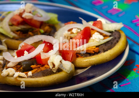 Mexikanische sopes gemacht mit Huhn tinga, Chilis, schwarz refried Beans und frische Zwiebel, Salat und Tomaten Stockfoto