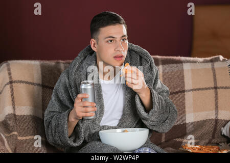 Hübscher junger Mann essen ungesundes Essen während der TV-Programmwiedergabe bei Nacht Stockfoto