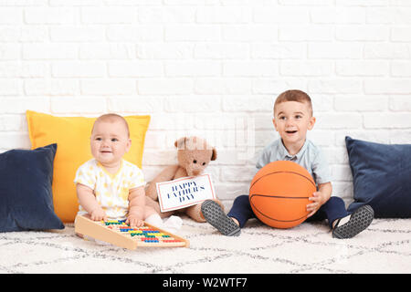 Süße kleine Kinder mit Spielzeug und Grußkarte für den Vatertag in der Nähe von White Brick Wall Stockfoto