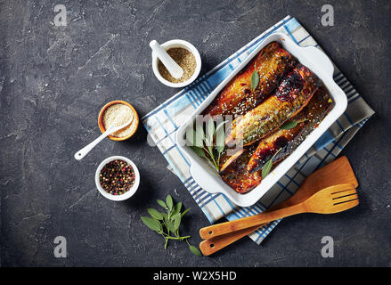 Lecker gebratene Makrelen in einer Auflaufform auf einer konkreten Tabelle mit Gewürzen im Hintergrund, Ansicht von oben, flach Stockfoto