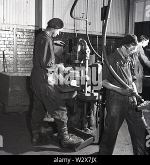 1950er Jahre, historische männliche Arbeiter in Overalls und Baskenmützen mit industriellen Werkzeugmaschinen auf dem Boden einer Batteriefabrik in Walthamstow, London, England, Großbritannien. Stockfoto