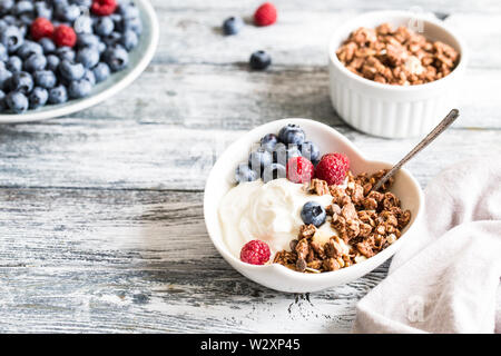 Griechischer Joghurt, Blaubeeren, Himbeeren und Müsli in einer weißen Schüssel auf einem hölzernen Hintergrund. Stockfoto