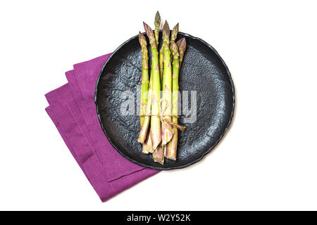 Frischer grüner Spargel auf schwarzer Keramik Teller und rosa Bettwäsche Serviette auf weißem Hintergrund, isoliert. Stockfoto
