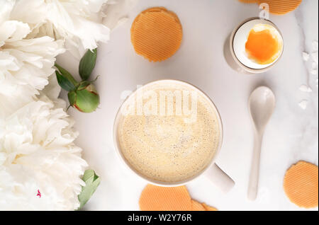 Blick von oben auf die weich gekochten Ei mit liquide orange Eigelb in Keramik Eierbecher, Tasse Kaffee mit Löffel aus Keramik, dünne knusprige Mais Chips und schöne weiße Pfingstrose. Frühstück Konzept. Flach. Stockfoto