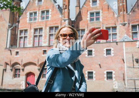 Stilvolle junge Frau Tourist in einer blauen Jacke macht einen selfie auf einem Mobiltelefon in Brügge, Belgien. Eine glückliche Frau steht vor dem Hintergrund der Stockfoto