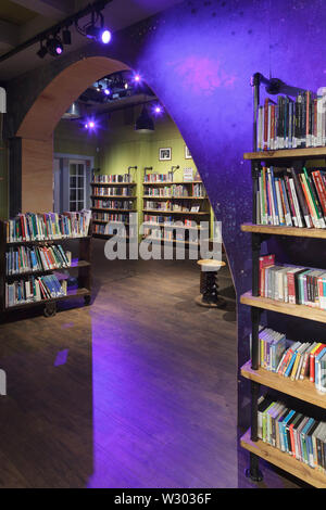 Lila spotlit wave-Form an der Wand" in der Kinderbibliothek, mit Bücherregalen. Bibliothek Deichman Grunerlokka, Oslo, Norwegen. Architekt: Aat Vos, 2019. Stockfoto