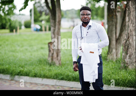 Junge afrikanische amerikanische männliche Arzt halten weißen Mantel an Hand mit einem Stethoskop outdoor aufgeworfen. Stockfoto