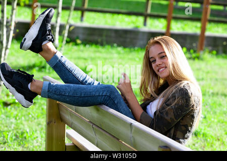 Spaß verspielte junge Frau sitzt auf einer Parkbank ihre Füße Schwingen in der Luft über den Rücken in die Kamera grinsend Stockfoto
