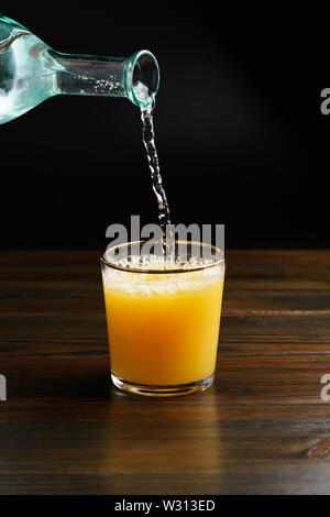 Der Dienst trinken, gießt Wasser in ein Glas mit Orangensaft. Dunkler Holztisch, hohe Auflösung Stockfoto