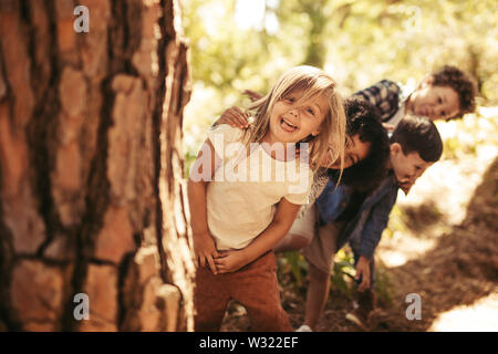 Gruppe von Kindern in der Zeile stehen und spähen hinter einem Baum im Freien. Kinder spielen verstecken und in einem Park suchen. Stockfoto