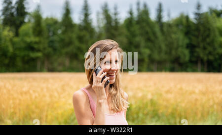 Junge Frau mit einem Lächeln auf ihrem Gesicht Gespräch am Handy, als sie einen Spaziergang in der Natur. Mit Weizenfeld im Hintergrund. Stockfoto