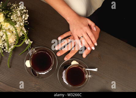 Bräutigam und Braut Hände mit Ringen auf dem Tisch mit zwei Kaffeetassen und Brautstrauß, Nahaufnahme der Ansicht von oben Stockfoto