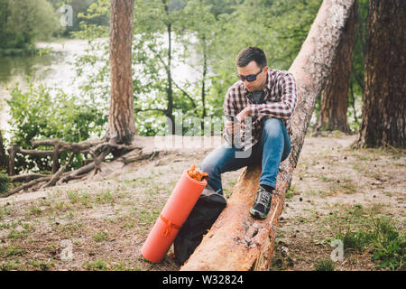 Junger Mann auf Lkw im Wald sitzen und über Handy. Mann Reisender auf großen gefallenen Baum sitzt, Telefon in der Hand halten. Thema Wandern Natur Reise Stockfoto