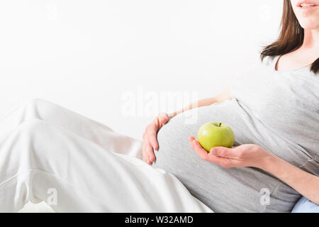 Schöne schwangere junge Frau mit dunklen Haaren tragen in leichter Kleidung und halten Grüner Apfel in der Hand. Diät Konzept. Kopieren Sie Platz. Gesunde Ernährung Stockfoto