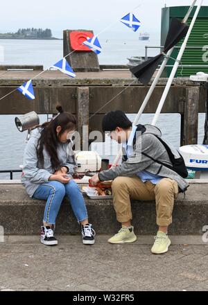 Asiatische Touristen teilen eine Platte mit Meeresfrüchten auf der Hafenmauer in Oban, Schottland, Großbritannien, Europa Stockfoto