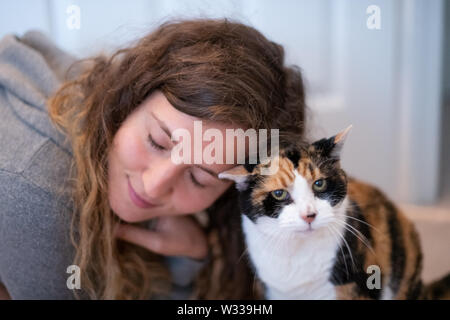 Closeup Portrait von Glücklich lächelnde junge Frau Verklebung mit Calico Cat pet Begleiter, Stoßen, Reiben bunting Köpfe, Freunde Zuneigung zeigen Stockfoto