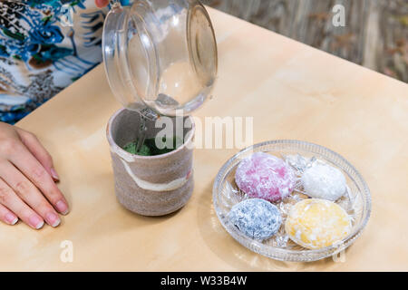 Frau gießen heißes Wasser in die Green Matcha Tee Tasse und mochi Reiskuchen japanische Dessert wagashi auf hölzernen Tisch Stockfoto