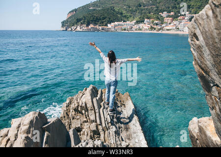 Ein Mädchen steht auf einem Felsen am Meer und hebt die Hände hoch. Sie schaut die schöne Aussicht auf die Küste mit der Küstenstadt Petrovac in Montenegro. Stockfoto