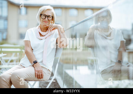 Lifestyle Portrait eines eleganten ältere Frau in Weiß sitzen auf der Terrasse des Cafés im Freien gekleidet. Konzept eines aktiven Lebensstils auf Ruhestand Stockfoto
