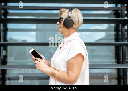 Lifwstyle Porträt einer stilvollen älteren Frau Anhören der Musik mit Kopfhörer und Telefon stehen auf dem modernen Gebäude Hintergrund Stockfoto