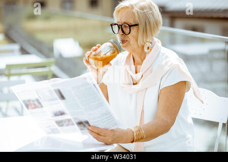 Elegante ältere Frau liest Zeitung, beim Sitzen auf der Terrasse des Cafés in der Stadt. Konzept eines aktiven Lebensstils auf Ruhestand Stockfoto