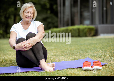 Porträt einer älteren Frau sitzt auf der Yogamatte, Sport im Park. Konzept eines aktiven Lebensstils auf Ruhestand Stockfoto