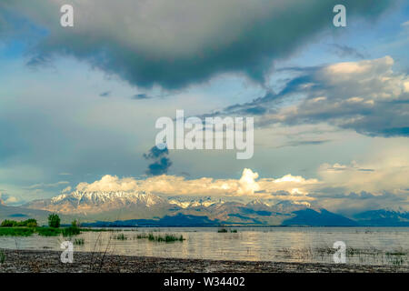 Malerische Ansicht der glänzenden See und schneebedeckten Berg bei bewölktem Himmel. Die helle Sonne ist hinter den dicken und geschwollene Wolken versteckt. Stockfoto