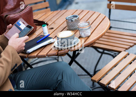 Freimütiger Moment eines Mannes, der aus der Ferne in einem Londoner Café mit Cappuccino, Smartphone und Notebook auf einem Holztisch arbeitet – urbanes Lifestyle-Konzept. Stockfoto