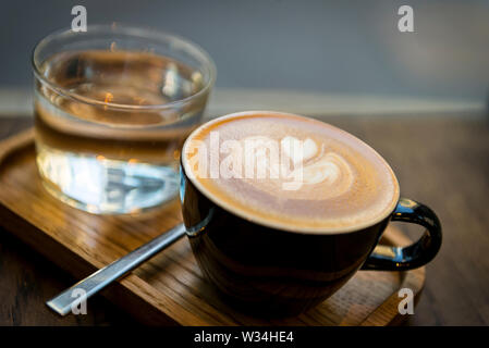 Latte Art in einer schwarzen Tasse auf einem Holztablett mit einem Glas Wasser und Löffel; gemütliches Café in der Nähe eines Espresso-Spezialgetränks für eine Kaffeepause am Morgen. Stockfoto