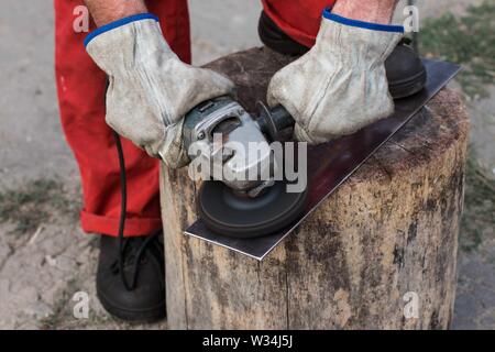 Der Master in einem roten Overall mit Händen in Arbeitshandschuhe schleift ein Stück Stahlblech mit einer alten Mühle Stockfoto