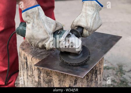 Eine industrielle Arbeiter in einer roten insgesamt mit seinen Händen in Arbeitshandschuhe schleift ein Stück Stahlblech mit einer alten Mühle Stockfoto
