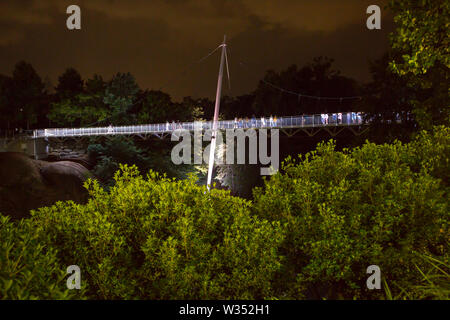 GREENVILLE, SC (USA) - Juli 5, 2019: Fußgänger überqueren Sie die hell erleuchtete Brücke in Falls Park bei Nacht. Stockfoto