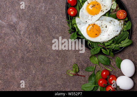 Spiegeleier mit Salat auf Pan auf dunklen braunen Hintergrund. Ansicht von oben, flach, freier Platz für Text Stockfoto