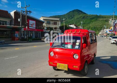 Ein rotes Tuk-tuk, ein Vierrädriges Taxi, befindet sich in der Hauptstraße durch Kamala, die geparkten, grünen Hügeln im b/g; Kamala Beach, Phuket, Thailand Stockfoto