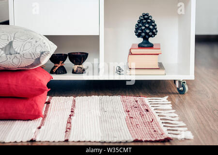 Gemütliche Einrichtung, weiße Nachttisch mit Stapel Bücher, Kerzen in Coconut Shell in der Nähe von einem bunten Teppich mit dekorativen Kissen Hintergrund Stockfoto