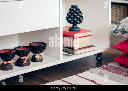 Gemütliche Einrichtung, weiße Nachttisch mit Stapel Bücher, Kerzen in Coconut Shell in der Nähe von einem bunten Teppich mit dekorativen Kissen Hintergrund Stockfoto