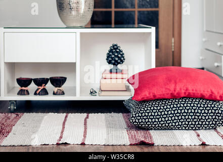 Gemütliche Einrichtung, weiße Nachttisch mit Stapel Bücher, Kerzen in Coconut Shell in der Nähe von einem bunten Teppich mit dekorativen Kissen Hintergrund Stockfoto