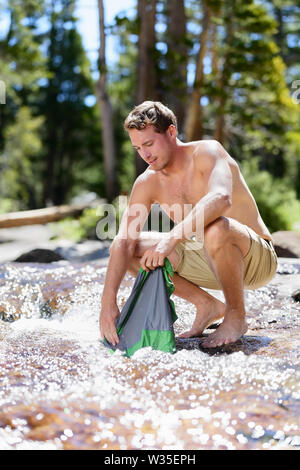 Camping Wandern Wanderer mann Wäsche waschen auf Wanderung in der Natur. Wandern junge männliche Erwachsene tun Kleidung waschen Chores in natürlichen Strom der frisches Wasser während einer Abenteuer Reise im Freien. Stockfoto