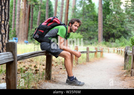 Wandern Menschen Porträt mit Rucksack müde Beine Ausruhen nach der langen Wanderung in der Natur. Kaukasische Mann lächelt zufrieden mit dem Wald im Hintergrund im Sommer Reise im Yosemite National Park, Kalifornien, USA. Stockfoto