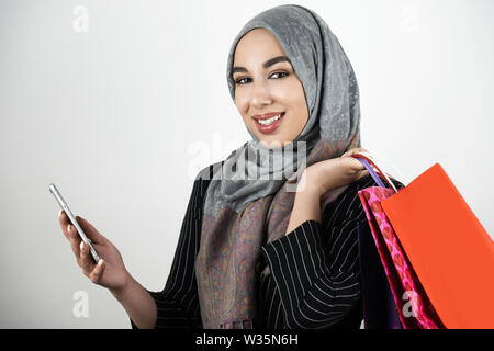 Junge schöne Lächeln Muslimischen business Frau mit Turban hijab Kopftuch holding Smartphone in der einen Hand und Einkaufstaschen in einem anderen isoliert Stockfoto