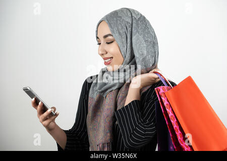 Junge schöne Lächeln Muslimischen business Frau mit Turban hijab Kopftuch Gewindebohren Smartphone mit einer Hand und Durchführung Einkaufstüten in einem anderen Stockfoto