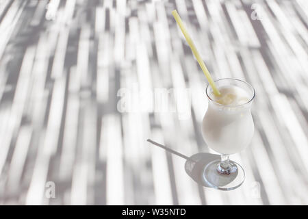 Pina Colada Cocktail mit gelben Trinkhalm, Kokosmilch und Eiswürfel auf weißen bar Tisch. Licht und Schatten spielen. Alkohol im Glas, summ Stockfoto