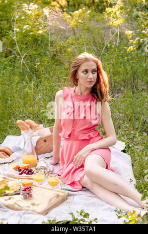 Die rothaarige Frau im Park. Picknick auf dem Rasen mit Korb, Brot, Käse und Obst Stockfoto