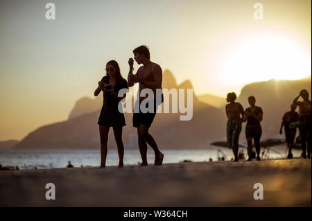 RIO DE JANEIRO - MÄRZ 20, 2018: die Menschen flanieren Sie entlang der Promenade in Arpoador Vergangenheit ein Sonnenuntergang Silhouette der beiden Brüder Berg am Strand von Ipanema. Stockfoto