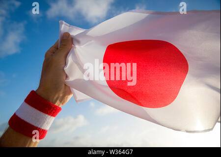 Hand mit Japan Rot und Weiß Armband mit einem japanischen Fahne winken in hellen, sonnigen blauen Himmel Stockfoto