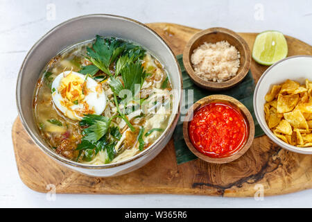 Soto Ayam, einem gelben scharfe Hühnersuppe mit gekochtem Ei, Petersilie und Bawang Goreng (gebratene Schalotten). Mit Kalk serviert, würzige sambal Sauce, Kartoffelchips Stockfoto