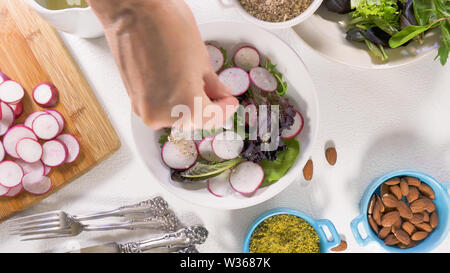 Kräutermischung Salat mit frischen organischen Radieschen, Kopfsalat, Spinat, einige Gewürze, Mandeln und Pflanzenöl. Nahaufnahme auf weißem Hintergrund, Frau Hände Stockfoto