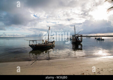 Kleine Segelboote auf dem Wasser am Strand bei Sonnenaufgang in Mafia Island, Tanzania, mit bewölktem Himmel und ruhiges Wasser mit umliegenden dugout Kanus. Stockfoto