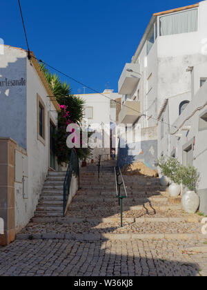 Suchen nach einem alten gepflasterten Straße mit Stufen und Handläufe mit einer Person zu Fuß unten zwischen den Reihenhäusern. Estoi, Portugal. Stockfoto